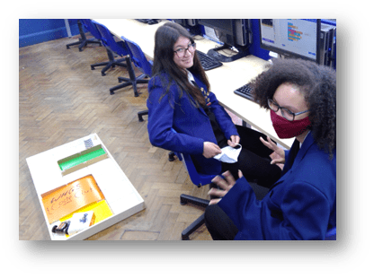 Two Year 9 girls are, sitting near a wooden rectangular arena containing a BitBot microbit buggy. They are in a classroom. One is smiling while wears a facemask but is making "jazz hands"!