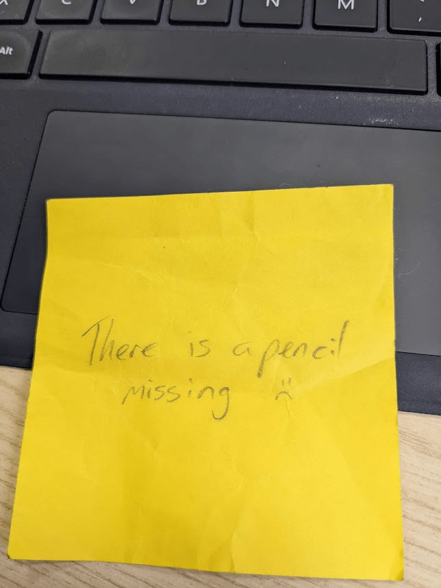 A yellow post-it note sits on a laptop keyboard with the words "There is a pencil missing" and a sad face emoji written on in pencil.