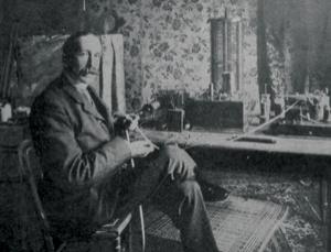 Neville Maskelyne sits in a chair in this black and white image from the 1900s. He's about 50 with receding hair and a moustache, sitting in a chair with some technical equipment nearby.